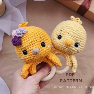 Könnte beinhalten: Zwei gehäkelte gelbe Kükenspielzeuge mit schwarzen Augen und Schnäbeln, die an einem Holz-Beißring befestigt sind. Das Küken links hat lila Blumen auf dem Kopf. Der Text "PDF PATTERN @HAPPINESS_BY_KRIS" ist unterhalb des Rings sichtbar.