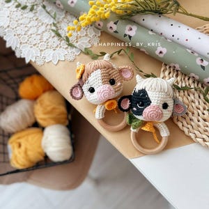 May include: Two crocheted cow rattle toys with wooden rings. One cow is brown and white with a yellow flower on its head. The other cow is black and white. The toys are on a brown surface with a white lace doily and a green and white patterned wrapping paper.