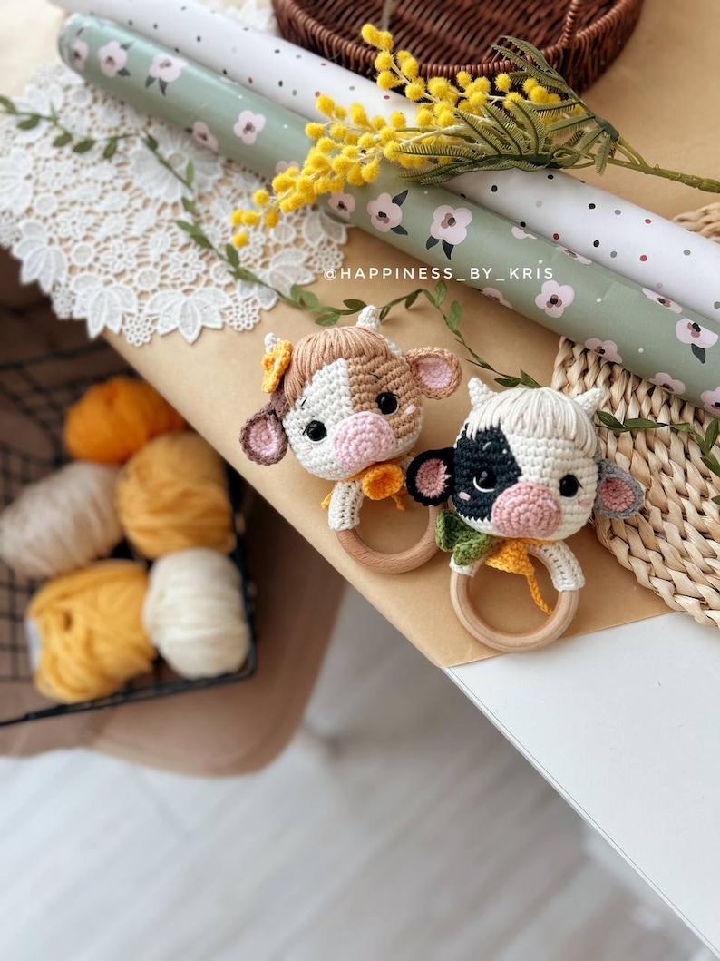 May include: Two crocheted cow rattle toys with wooden rings. One cow is brown and white with a yellow flower on its head. The other cow is black and white. The toys are on a brown surface with a white lace doily and a green and white patterned wrapping paper.