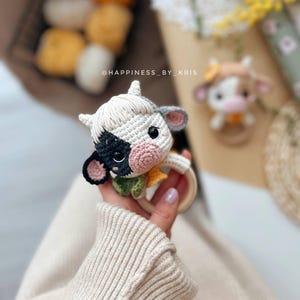 May include: A crocheted black and white cow rattle toy with a wooden ring. The toy is held in a hand with a white sweater.