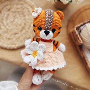 May include: A crocheted tiger plush toy wearing a peach-colored dress with a white daisy on the front. The tiger has orange and black stripes and a white face with black eyes and a black nose. The toy is holding a white daisy in its paw. The text "@HAPPINESS_BY_KRIS" is visible in the bottom right corner of the image.