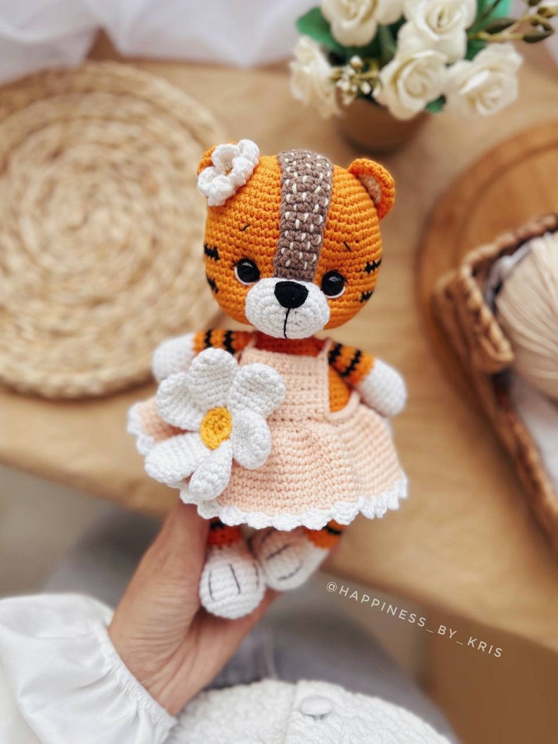 May include: A crocheted tiger plush toy wearing a peach-colored dress with a white daisy on the front. The tiger has orange and black stripes and a white face with black eyes and a black nose. The toy is holding a white daisy in its paw. The text "@HAPPINESS_BY_KRIS" is visible in the bottom right corner of the image.