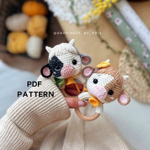 May include: Two crocheted cow plushies with wooden rings, one black and white and one brown and white, with the text "PDF PATTERN" and "@HAPPINESS_BY_KRIS" on the image.