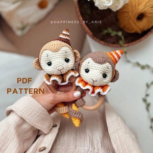 以下が含まれることがあります： オレンジと黄色のアクセントが付いた、2つの編みぐるみのおさるの玩具が、木製のおしゃぶりリングを持っています。おもちゃはパーティーハットをかぶり、首にフリルが付いています。画像には「PDF PATTERN」というテキストが表示されています。