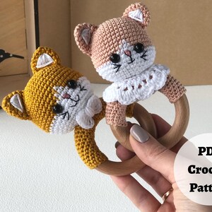 May include: Two crocheted cat rattle toys with wooden rings. One cat is yellow and the other is peach.