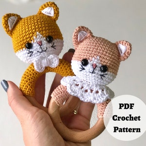 May include: Two crocheted cat rattle toys with wooden rings. One is yellow and the other is pink. Both have white crocheted details and black eyes.
