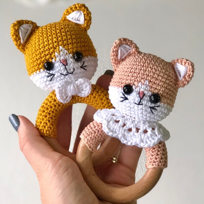 May include: Two crocheted cat rattle toys with wooden rings. One is yellow and the other is pink.
