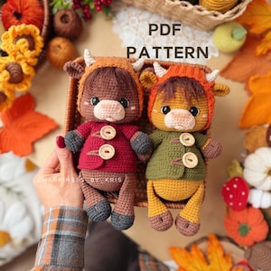 Könnte beinhalten: Zwei gehäkelte Kuhpuppen mit Hörnern, eine in einer dunkelroten Jacke und die andere in einer grünen Jacke, beide mit Holzknebelknöpfen. Der Text PDF PATTERN ist über den Puppen in einem geflochtenen Korb zu sehen.