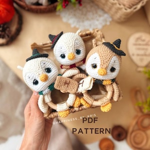 May include: Three crocheted penguin toys with black hats, each holding a wooden ring. The penguins have white and tan bodies, yellow beaks, and are wearing scarves. The text "PDF PATTERN" is visible.