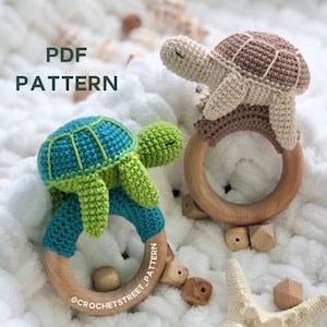 Puede incluir: Dos sonajeros de bebé de tortuga de ganchillo. Una tortuga es turquesa y verde, la otra es beige y marrón. Cada tortuga está unida a un anillo de madera. El texto "PDF PATTERN" está en la esquina superior izquierda. El texto "@crochetstreet_pattern" está en el anillo de madera.