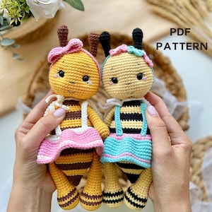 Può includere: Due bambole a forma di ape fatte a mano all'uncinetto. Una bambola a forma di ape ha un fiocco e una gonna rosa, l'altra una gonna blu. Entrambe hanno corpi a strisce nere e gialle e la scritta "PDF PATTERN". Le bambole sono tenute da una persona.