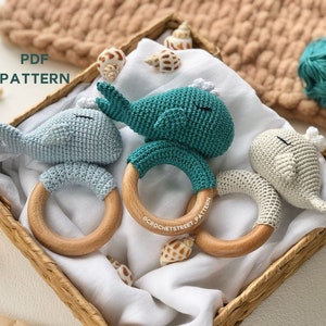 May include: Three crocheted whale teethers in blue, green, and gray colors attached to wooden rings. The teethers are lying on a white fabric surface inside a wicker basket. The text "PDF PATTERN" is visible in the top left corner of the image.