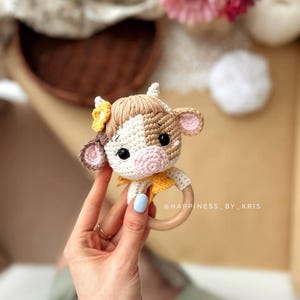 May include: A crocheted cow rattle toy with brown, white, and pink accents. The rattle has a wooden ring and a yellow flower on its head. The toy is held in a hand with blue nail polish.