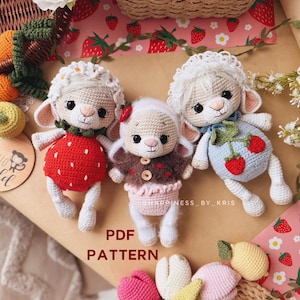 Puede incluir: Tres muñecos de cordero tejidos a mano con hilo color crema, grandes ojos negros y atuendos distintos. Uno lleva un vestido rojo con estampado de fresas, otro una chaqueta marrón con falda rosa y el tercero un vestido azul claro con fresas.