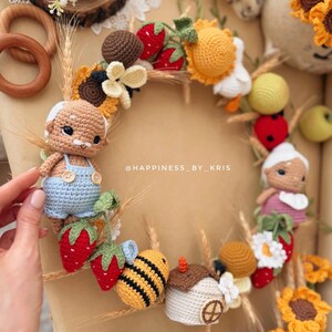 K&ouml;nnte beinhalten: Ein runder H&auml;kelkranz mit zwei kleinen Puppen, einer Biene, einem Haus, Erdbeeren, Sonnenblumen und Weizen&auml;hren. Der Kranz ist mit verschiedenen Elementen aus Garn verziert und enth&auml;lt den Text @HAPPINESS_BY_KRIS in der Mitte.