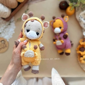 May include: Two handmade crochet giraffe dolls. One is held in a hand and is wearing a yellow jumpsuit with brown spots and accents. The other is wearing a purple jumpsuit. Both have giraffe-like features.