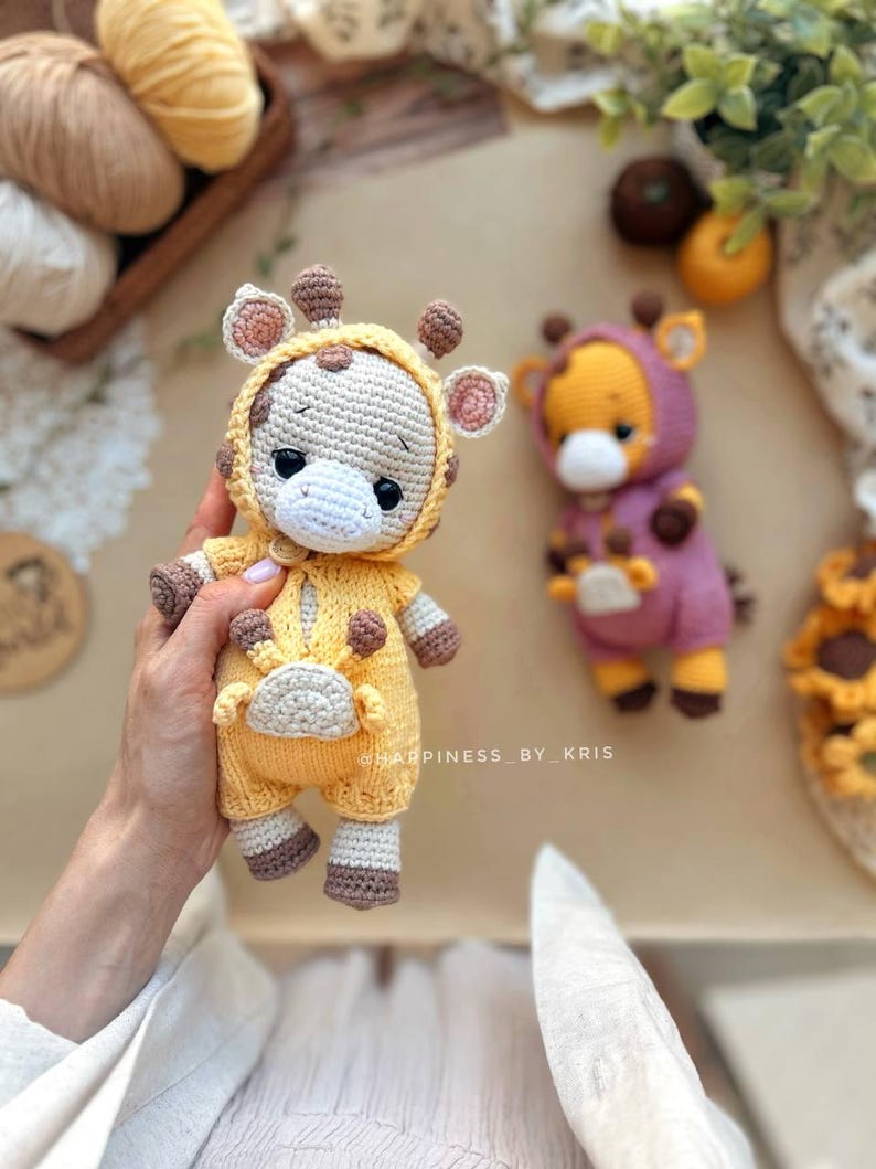 May include: Two handmade crochet giraffe dolls. One is held in a hand and is wearing a yellow jumpsuit with brown spots and accents. The other is wearing a purple jumpsuit. Both have giraffe-like features.