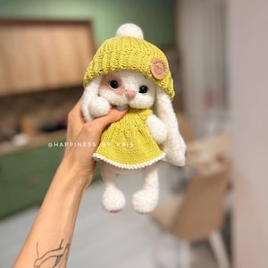 May include: A white crocheted bunny wearing a yellow knitted hat and dress. The bunny has a small, white pom-pom on top of its hat and a brown tag that says "handmade" with a heart.