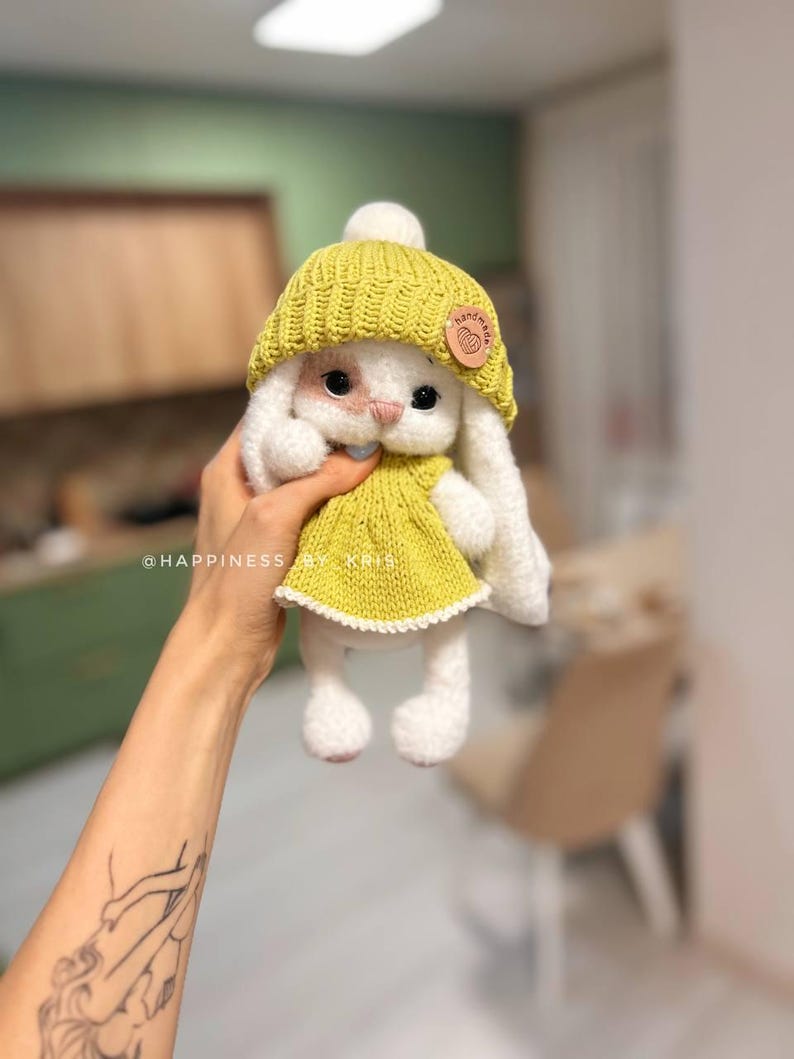 May include: A white crocheted bunny wearing a yellow knitted hat and dress. The bunny has a small, white pom-pom on top of its hat and a brown tag that says "handmade" with a heart.