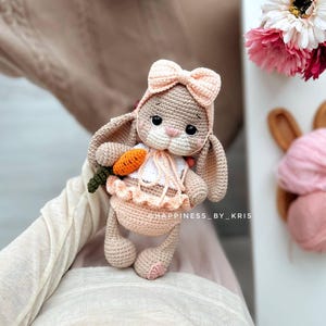 May include: A crocheted bunny toy wearing a pink bow and holding a carrot. The bunny is sitting on a person's lap. The toy is made with soft yarn and has a sweet expression.