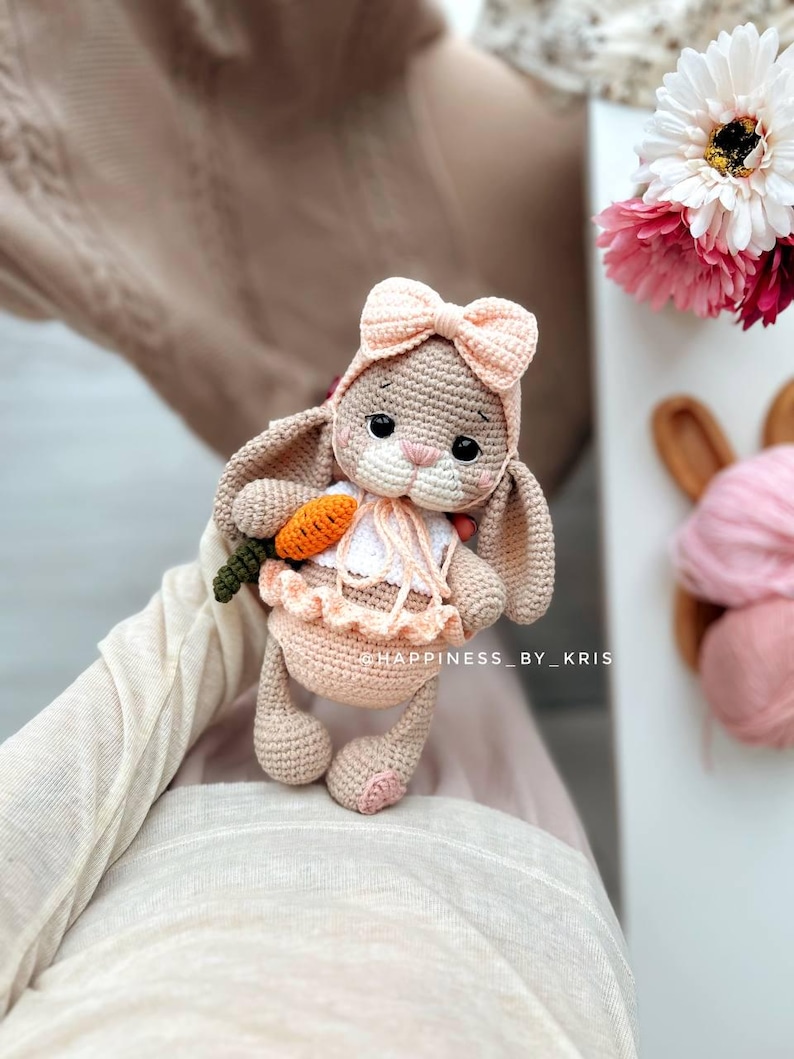 May include: A crocheted bunny toy wearing a pink bow and holding a carrot. The bunny is sitting on a person's lap. The toy is made with soft yarn and has a sweet expression.