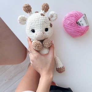 May include: A crocheted giraffe toy with brown spots and a pink yarn ball. The giraffe is white with brown accents and has a sweet expression.