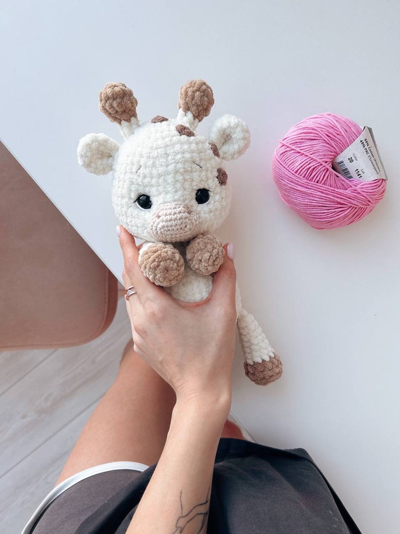 May include: A crocheted giraffe toy with brown spots and a pink yarn ball. The giraffe is white with brown accents and has a sweet expression.