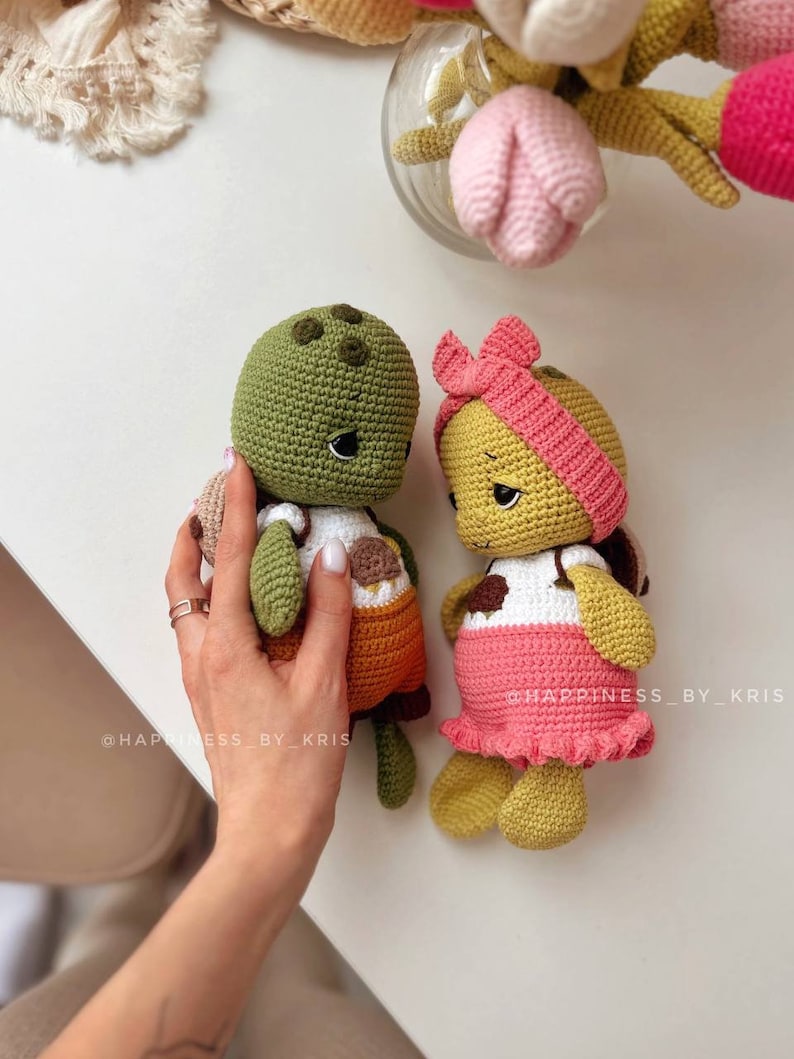 May include: Two crocheted turtle plushies, one green with orange accents and the other yellow with pink accents. Both have black eyes and smiling expressions. The turtles are sitting on a white surface. The text "@happiness_by_kris" is visible on the bottom of the image.