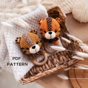 Könnte beinhalten: Zwei gehäkelte Tiger-Rasselspielzeuge mit braunen und orangen Streifen, einem Holzring und einer weißen Schleife. Die Spielzeuge befinden sich in einem Weidenkorb mit weißen, flauschigen Stielen.