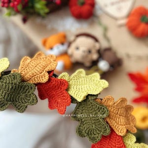 K&ouml;nnte beinhalten: Nahaufnahme einer geh&auml;kelten Bl&auml;ttergirlande in Herbstfarben: Gr&uuml;n, Orange, Gelb und Braun. Die Girlande ist im Fokus, mit einem verschwommenen Hintergrund mit kleinen K&uuml;rbissen, Beeren und geh&auml;kelten Tieren. Der Text "@HAPPINESS BY KRIS" ist sichtbar.