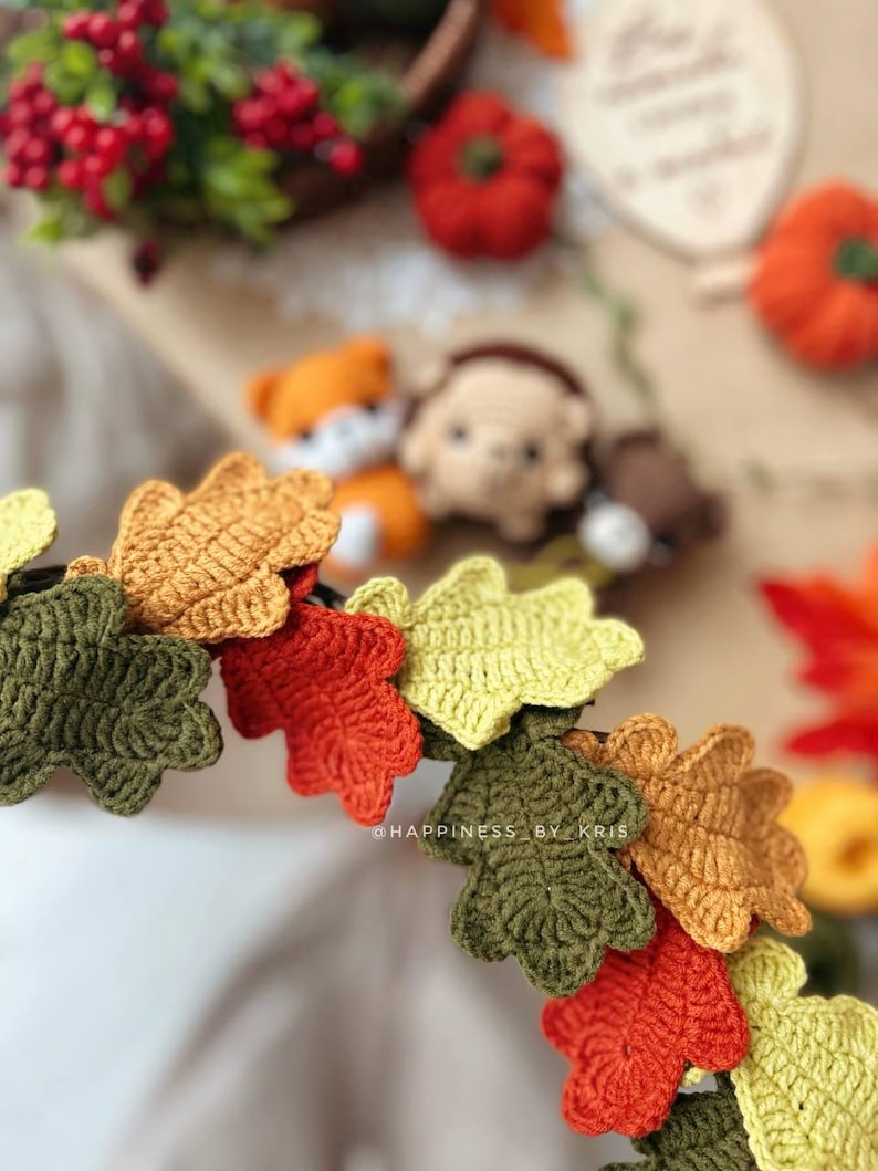 K&ouml;nnte beinhalten: Nahaufnahme einer geh&auml;kelten Bl&auml;ttergirlande in Herbstfarben: Gr&uuml;n, Orange, Gelb und Braun. Die Girlande ist im Fokus, mit einem verschwommenen Hintergrund mit kleinen K&uuml;rbissen, Beeren und geh&auml;kelten Tieren. Der Text "@HAPPINESS BY KRIS" ist sichtbar.
