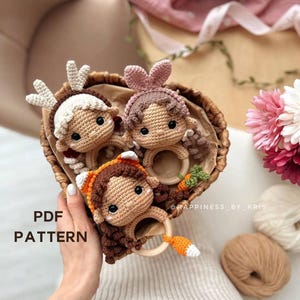 Könnte beinhalten: Drei gehäkelte Babyspielzeuge in einem Weidenkorb. Die Spielzeuge sind ein Reh, ein Kaninchen und ein Fuchs. Jedes Spielzeug hat einen Holzring daran befestigt. Die Spielzeuge sind braun, beige und orange. Der Korb ist braun und hat eine weiße Auskleidung. Der Text "PDF PATTERN" ist im Bild sichtbar.