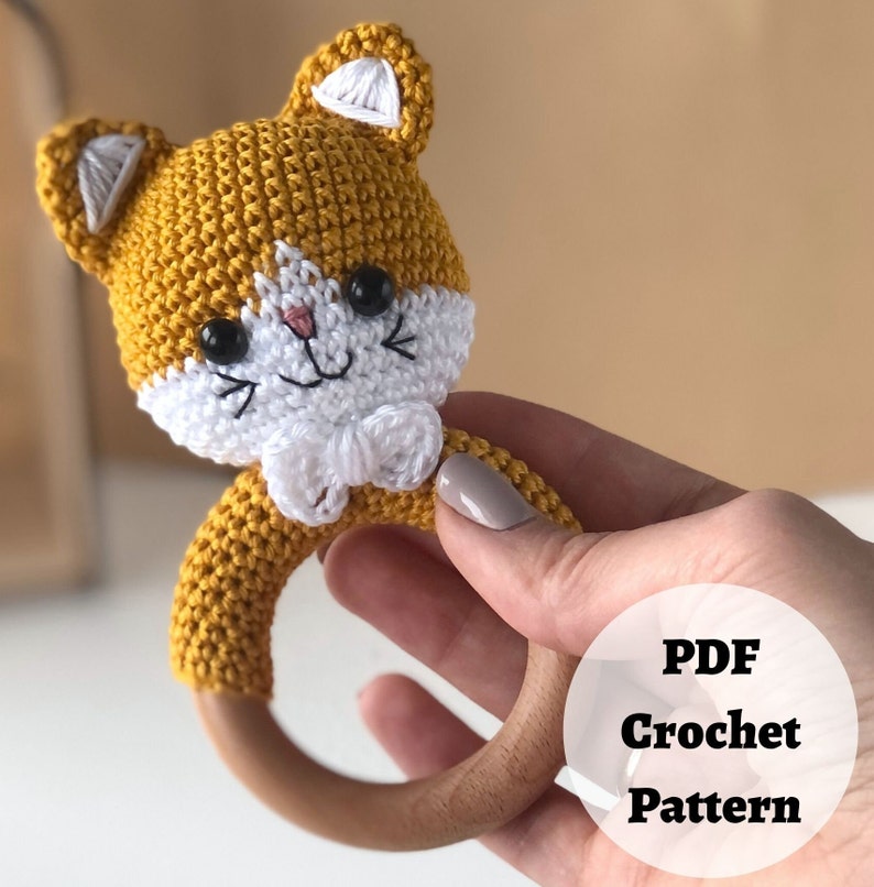 May include: Crochet pattern for a yellow and white cat rattle toy. The rattle is made of crocheted yarn and has a wooden ring. The pattern is available as a PDF download.