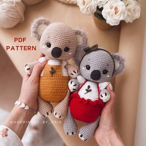 May include: Two crocheted koala amigurumi toys. One is beige with a yellow and white outfit and the other is grey with a red and white outfit. The toys are being held by a person's hands. The text "PDF PATTERN" is visible in the image.