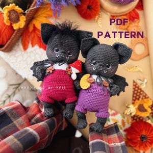 May include: Two black crocheted bat dolls with large ears and wings. One wears red overalls with a mushroom, the other purple overalls with a yellow moon. Both have white fangs and dark eyes. Text reads PDF PATTERN and HAPPINESS BY KRIS.