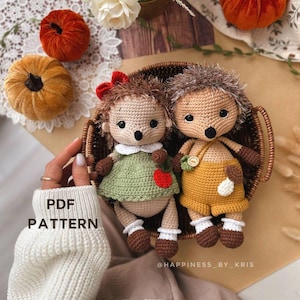 Könnte beinhalten: Zwei gehäkelte Igel-Amigurumi-Spielzeuge in einem Weidenkorb. Der Igel links trägt ein grünes Kleid mit einem roten Apfel und einer roten Schleife. Der Igel rechts trägt eine gelbe Latzhose mit einem weißen Knopf und einer weißen Blume. Der Text "PDF PATTERN" ist im Bild sichtbar.