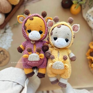 May include: Two handmade crochet giraffe dolls. One is yellow and purple, the other is yellow and cream. Both have brown spots, ears, and horns. They are wearing hooded onesies with button closures. The text "@HAPPINESS BY KRIS" is visible.