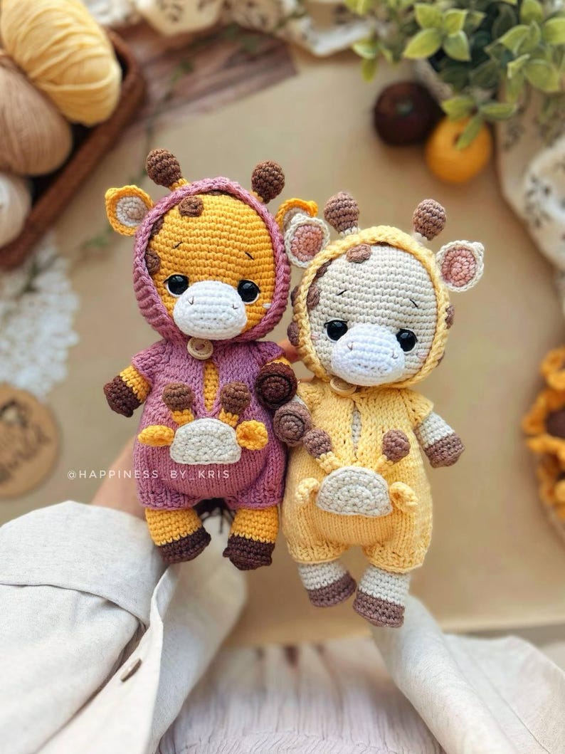 May include: Two handmade crochet giraffe dolls. One is yellow and purple, the other is yellow and cream. Both have brown spots, ears, and horns. They are wearing hooded onesies with button closures. The text "@HAPPINESS BY KRIS" is visible.