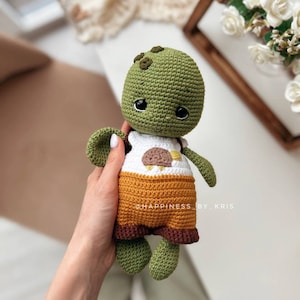 May include: A green crocheted turtle wearing a brown and orange outfit with a white shirt featuring a brown bird design. The turtle has black eyes and a small smile. The text "@HAPPINESS_BY_KRIS" is visible in the image.