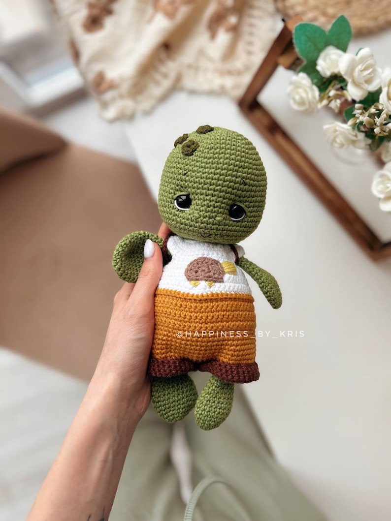 May include: A green crocheted turtle wearing a brown and orange outfit with a white shirt featuring a brown bird design. The turtle has black eyes and a small smile. The text "@HAPPINESS_BY_KRIS" is visible in the image.
