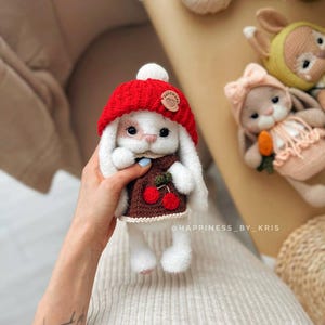 May include: White crocheted bunny wearing a red knitted hat with a white pom pom and a brown knitted sweater with cherries. The bunny is holding a small green stem with a cherry on it. The bunny is sitting on a person's hand.