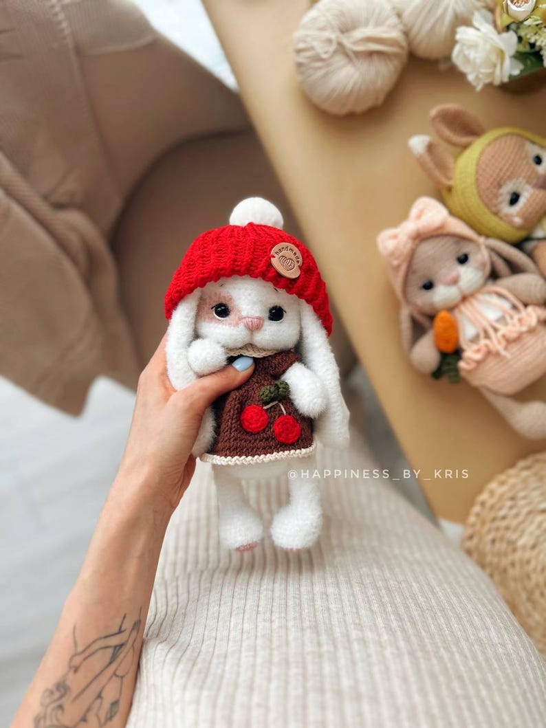 May include: White crocheted bunny wearing a red knitted hat with a white pom pom and a brown knitted sweater with cherries. The bunny is holding a small green stem with a cherry on it. The bunny is sitting on a person's hand.