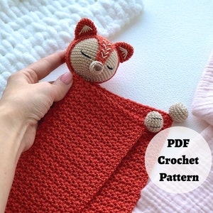 Peut inclure: Une couverture de s&eacute;curit&eacute; en crochet en forme de renard avec une couverture rouge et une t&ecirc;te de renard beige. La couverture est dot&eacute;e de deux boules en crochet beige attach&eacute;es &agrave; elle. Le texte "PDF Crochet Pattern" est visible sur l'image.