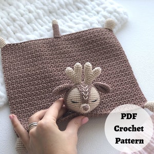 Peut inclure: Une couverture b&eacute;b&eacute; en crochet marron avec une applique de cerf en crochet. La couverture a des franges sur les bords et le cerf a un visage cousu. Le texte "PDF Crochet Pattern" est visible sur l'image.