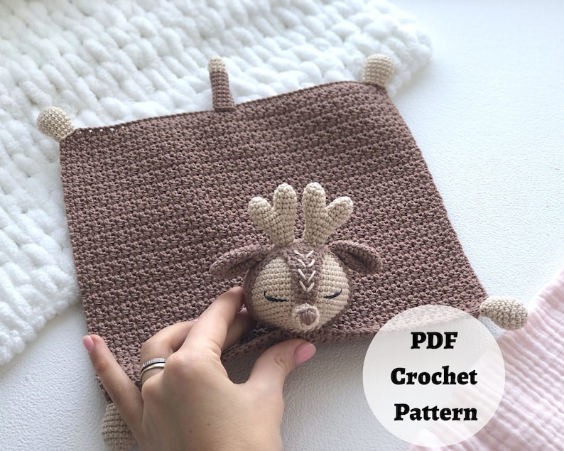 Peut inclure: Une couverture b&eacute;b&eacute; en crochet marron avec une applique de cerf en crochet. La couverture a des franges sur les bords et le cerf a un visage cousu. Le texte "PDF Crochet Pattern" est visible sur l'image.