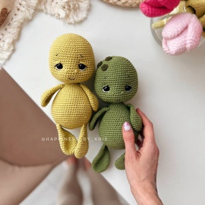 May include: Two crocheted stuffed toys, one yellow and one green, with embroidered eyes and smiling expressions. The toys are sitting on a white surface. The text "@HAPPINESS_BY_KRIS" is visible in the lower left corner of the image.