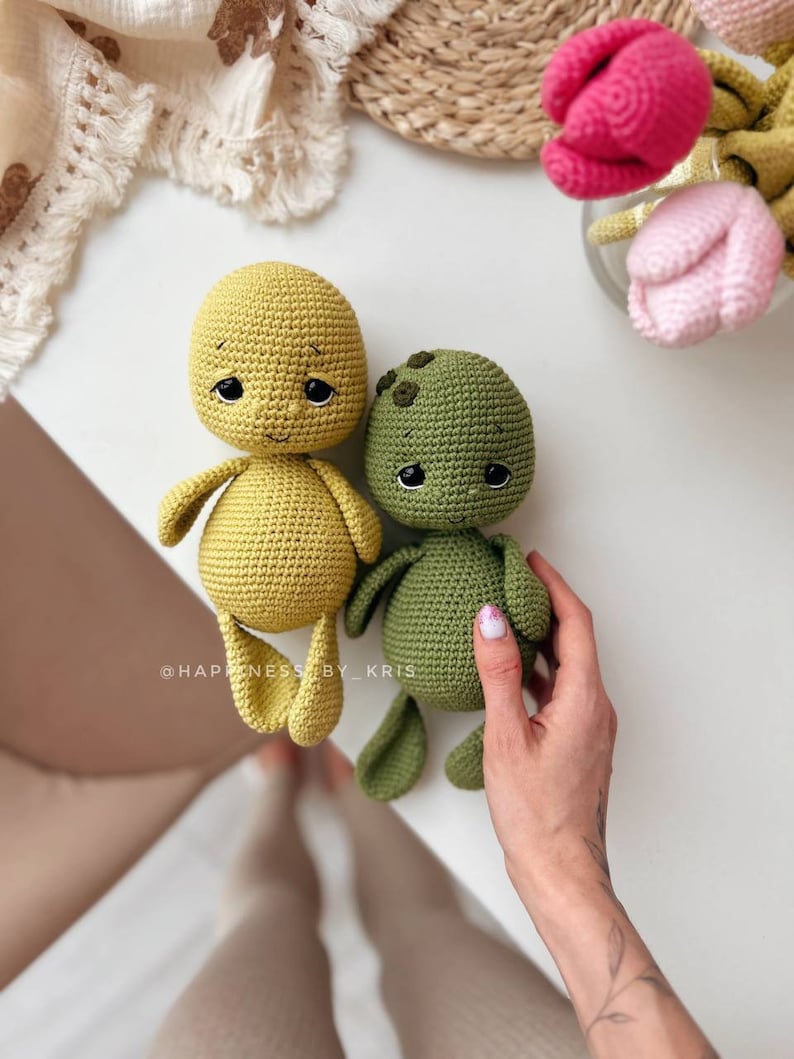 May include: Two crocheted stuffed toys, one yellow and one green, with embroidered eyes and smiling expressions. The toys are sitting on a white surface. The text "@HAPPINESS_BY_KRIS" is visible in the lower left corner of the image.