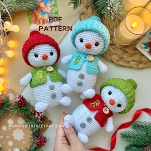Puede incluir: Tres muñecos de nieve de ganchillo con gorros y chalecos rojos, verdes y azules. Cada muñeco de nieve tiene una nariz de zanahoria, ojos de botón y un botón en el chaleco. La imagen incluye el texto "PDF PATTERN" y el identificador de Instagram "@CROCHETSTREETPATTERN".