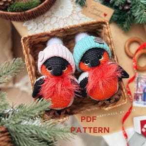 May include: Two crocheted bird ornaments with orange bodies, black heads, and fluffy red chests, wearing pink and blue knitted hats with white bobbles. They sit in a wicker basket, with the text "PDF PATTERN" visible.