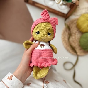May include: A crocheted doll with a pink dress and headband. The doll has a yellow body and a white shirt with a brown turtle design. The doll is being held by a hand.